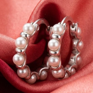 May include: A pair of silver-toned hoop earrings featuring a series of small, round, white pearls embedded along the outer edge of each circular band. The earrings have a hinged closure mechanism and a polished metallic finish on the silver-toned metal.