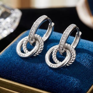 May include: A pair of silver hoop earrings featuring a hinged closure and a row of small clear stones along the outer edge. Each earring includes a dangling, textured rope-style circular charm engraved with the text 925 on the bottom surface.