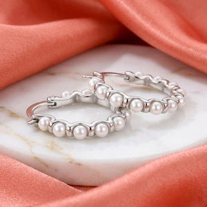 Può includere: Un paio di orecchini a cerchio in argento caratterizzati da una serie di piccole perle bianche rotonde incastonate lungo il bordo esterno di ogni fascia circolare. Gli orecchini sono realizzati in materiale metallico lucido con chiusura.