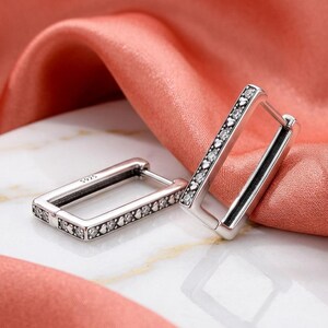 May include: A pair of rectangular hoop earrings crafted from silver-toned metal, each featuring a row of small, clear, sparkling stones and tiny heart-shaped cutouts along the outer edge. The inner side of the hoop is stamped with S925.