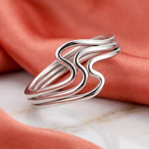 May include: A polished silver-toned ring featuring a multi-layered design with three distinct, smooth, wavy bands that curve and overlap to create a continuous, fluid, and interconnected shape that wraps around the finger.