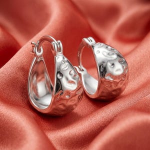 May include: A pair of silver hoop earrings featuring a textured, hammered surface finish. Each earring is constructed with a hinged post mechanism and displays a small 925 hallmark stamp on the side, indicating sterling silver composition.