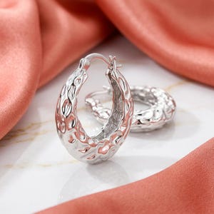 May include: A pair of silver-toned hoop earrings featuring a textured, hammered surface with an irregular, organic pattern. The hoops are oval-shaped and include a hinged post closure mechanism, showcasing a reflective metallic finish throughout.