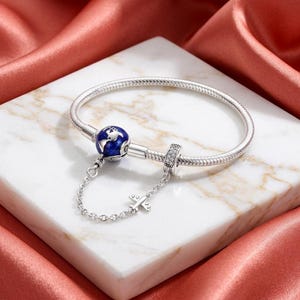 May include: A silver snake chain bracelet featuring a spherical charm with a blue enamel world map design, a small silver aeroplane charm attached by a delicate chain, and a cylindrical spacer bead encrusted with clear stones.
