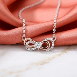 May include: A necklace featuring a silver-toned chain and a central infinity-shaped pendant. The pendant combines smooth rose-gold-toned metal with twisted silver-toned metal, centred with a small, clear, heart-shaped faceted gemstone.