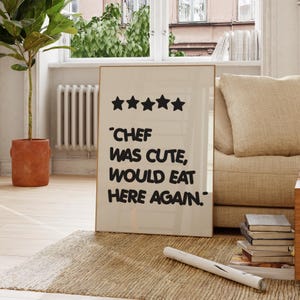 Op de afbeelding: Een ingelijste print met de tekst "CHEF WAS CUTE, WOULD EAT HERE AGAIN" en vijf zwarte sterren. De print staat in een kamer met een beige tapijt, een beige bank en een plant in een pot.