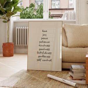 Op de afbeelding: Een ingelijste print met een gouden rand toont een lijst met woorden in zwart schrift: liefde, vreugde, vrede, geduld, vriendelijkheid, goedheid, trouw, zachtmoedigheid en zelfbeheersing. De print leunt tegen een beige bank en boeken.