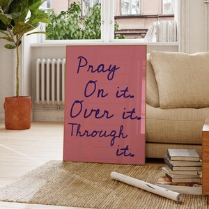 Op de afbeelding: Een ingelijste roze print met de woorden "Pray On it. Over it. Through it." in donkerblauw script. De print wordt weergegeven op een tapijt voor een raam en een beige bank.