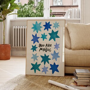 Op de afbeelding: Een ingelijste kunstprint met blauwe en turquoise sterren op een crèmekleurige achtergrond. De tekst "YOU ARE MAGIC" staat in het zwart. Het kunstwerk zit in een gouden lijst, ideaal voor interieurdecoratie, ongeveer 50 cm x 70 cm.