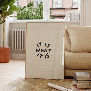Op de afbeelding: Een ingelijste kunstafdruk met de zin "IT IS WHAT IT IS" in zwart handschrift. Het kunstwerk wordt tentoongesteld in een lichte kamer met een raam, een plant en een beige bank.