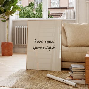 Op de afbeelding: Een ingelijste print met de woorden "love you goodnight" in zwart schrift. De print heeft een crèmekleurige achtergrond en een dunne gouden lijst. De print leunt tegen een beige bank in een goed verlichte kamer.