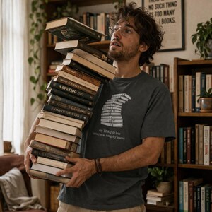 May include: A person holding a large stack of books, with titles visible like "Sapiens" and "Name of the Wind." The person wears a dark gray t-shirt with a graphic of stacked books and the text "my TBR pile has structural integrity issues."