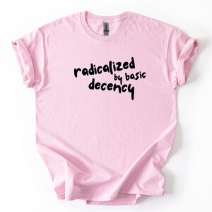 Könnte beinhalten: Hellrosa T-Shirt mit dem schwarzen Schriftzug "radicalized by basic decency" in handschriftlicher Schrift. Das T-Shirt hat hochgekrempelte Ärmel. Der Hintergrund ist weiß.
