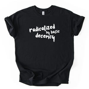 Könnte beinhalten: Schwarzes T-Shirt mit dem weißen Text "radicalized by basic decency". Das T-Shirt ist gefaltet und liegt flach auf weißem Hintergrund. Der Text ist in einer handschriftlichen Schriftart.