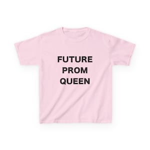 Op de afbeelding: Een lichtroze T-shirt met de zwarte tekst "FUTURE PROM QUEEN" in blokletters. Het shirt heeft korte mouwen en een ronde hals, geschikt voor casual wear.