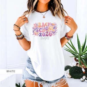 Puede incluir: Una camiseta blanca de manga corta con un estampado gráfico colorido con el texto "BACK SCHOOL 2026" y un espacio para un nombre. El diseño incluye salpicaduras de estilo acuarela en morado, naranja y rosa, estrellas e iconos escolares.