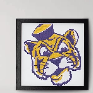 May include: Framed cross-stitch artwork depicting a tiger's head in yellow and purple. The tiger wears a hat. The piece is set against a white background and enclosed in a black square frame.
