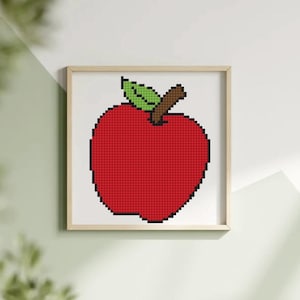 Puede incluir: Impresión enmarcada de pixel art de una manzana roja con una hoja verde y un tallo marrón. La manzana está compuesta por pequeños cuadrados rojos, lo que le da una estética retro y digital. La obra de arte está enmarcada en un marco de madera clara.