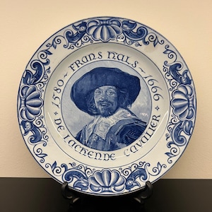 May include: A decorative ceramic plate featuring a portrait of Frans Hals, a Dutch Golden Age painter. The plate has a blue and white color scheme with ornate floral and scrollwork borders. Text includes "Frans Hals" and dates 1580-1666.