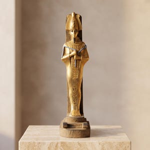 Puede incluir: Una estatua dorada de un faraón egipcio con una corona alta, sosteniendo un cayado y un flagelo azules. La figura se alza contra un panel trasero vertical oscuro, con jeroglíficos detallados tallados en el torso y las piernas.