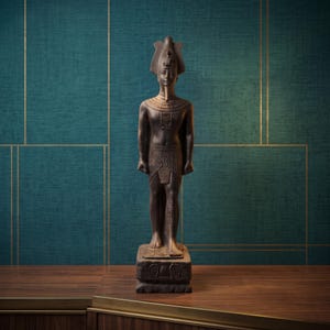 Puede incluir: Una estatua de pie de color marrón oscuro de un faraón egipcio con un tocado nemes tradicional y un faldellín plisado. La figura presenta un collar ancho, una barba ceremonial y se apoya sobre una base rectangular decorada con jeroglíficos.