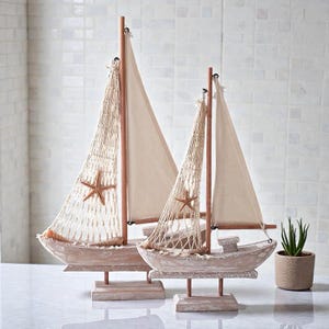 Puede incluir: Dos veleros decorativos de madera con velas color crema y redes. Cada barco presenta una estrella de mar y se apoya en un soporte de madera. Los barcos están pintados con un acabado blanco envejecido, evocando un tema náutico.