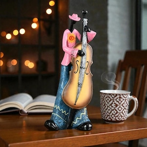 May include: A decorative figurine of a musician playing a double bass. The figure wears a pink shirt with a sun design, blue trousers, and a pink hat. The double bass is yellow and brown. A mug of coffee sits nearby.