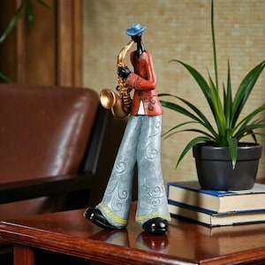 May include: A decorative figurine of a jazz musician playing a saxophone. The figure is dressed in a red jacket, blue trousers, and a blue hat. The saxophone is gold-coloured, and the figure has black skin.