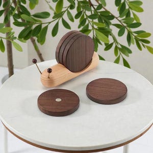 Puede incluir: Un juego de posavasos de madera con forma de caracol. El cuerpo del caracol es de madera de color claro, y la concha está hecha de posavasos de color marrón oscuro. Dos posavasos adicionales están junto al caracol. El caracol tiene dos pequeñas antenas con puntas rojas.