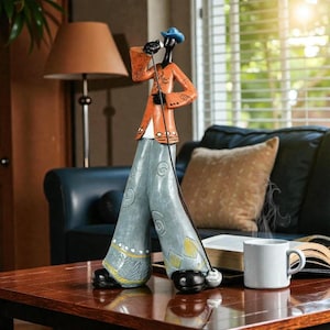 May include: A decorative figurine of a singer in a blue hat and orange jacket, holding a microphone. The figurine has grey trousers with decorative swirls and yellow accents. The singer is standing on a table with a white mug.