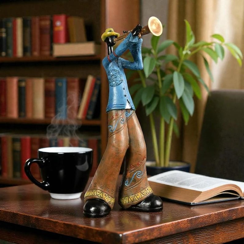 May include: A decorative figurine of a jazz musician playing a trumpet. The figure wears a blue jacket, brown trousers, and a yellow hat. A black coffee mug and an open book are on the table.