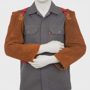 May include: A grey button-down shirt with two chest pockets and a small red tag, worn with brown leather protective sleeves. The sleeves are secured by red straps that cross over the shoulders and feature a small gold geometric logo design.