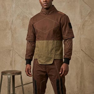 May include: Brown two-piece outfit with geometric stitching and accents. The top has a high collar, short sleeves with patterned fabric, and a contrasting olive green panel. The pants have black panels and stitching details. A metal stool is in the background.