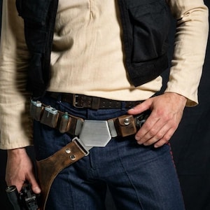 Op de afbeelding: Een beige shirt met lange mouwen, een zwart vest en een donkerblauwe broek gedragen met een bruine leren utility-riem. De riem heeft een grote metalen zeshoekige gesp, verschillende kleine tasjes en een holster voor een wapen.
