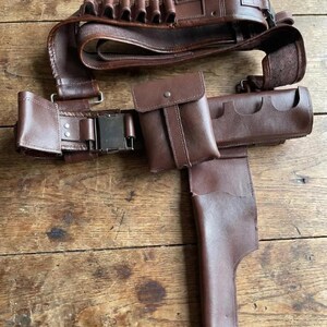 May include: A brown leather gun belt with multiple cartridge holders, a pouch, and a holster. The belt has a metal buckle and is designed to be worn around the waist. The leather has a worn appearance.