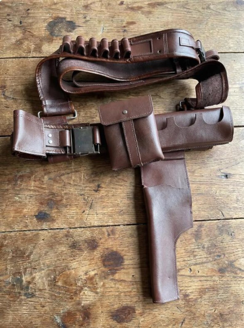 May include: A brown leather gun belt with multiple cartridge holders, a pouch, and a holster. The belt has a metal buckle and is designed to be worn around the waist. The leather has a worn appearance.