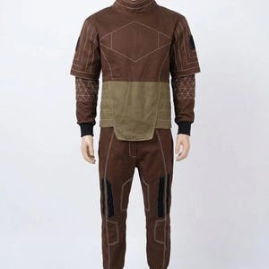 May include: A brown two-piece outfit on a mannequin. The top has a high collar and geometric stitching. The midsection is olive green. The pants have black accents on the knees and black cuffs. The outfit is designed with a futuristic aesthetic.