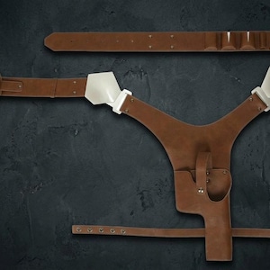 Op de afbeelding: Een bruin lederen schouderholstersysteem met een Y-vormig harnas met witte metalen schouderplaten, een gevormde pistoolholster en een verstelbare riem met metalen hardware. Een bijpassende bruine lederen riem is inbegrepen.