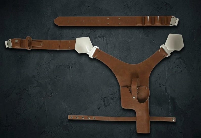Op de afbeelding: Een bruin lederen schouderholstersysteem met een Y-vormig harnas met witte metalen schouderplaten, een gevormde pistoolholster en een verstelbare riem met metalen hardware. Een bijpassende bruine lederen riem is inbegrepen.