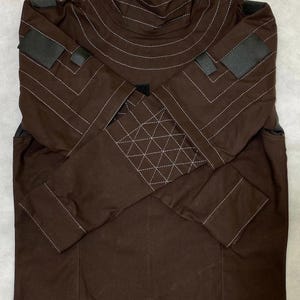 Puede incluir: Chaqueta marr&oacute;n con cuello alto y mangas largas. La chaqueta presenta detalles de costura blancos y parches negros en los hombros y las mangas. Una cremallera recorre la parte delantera del cuello.