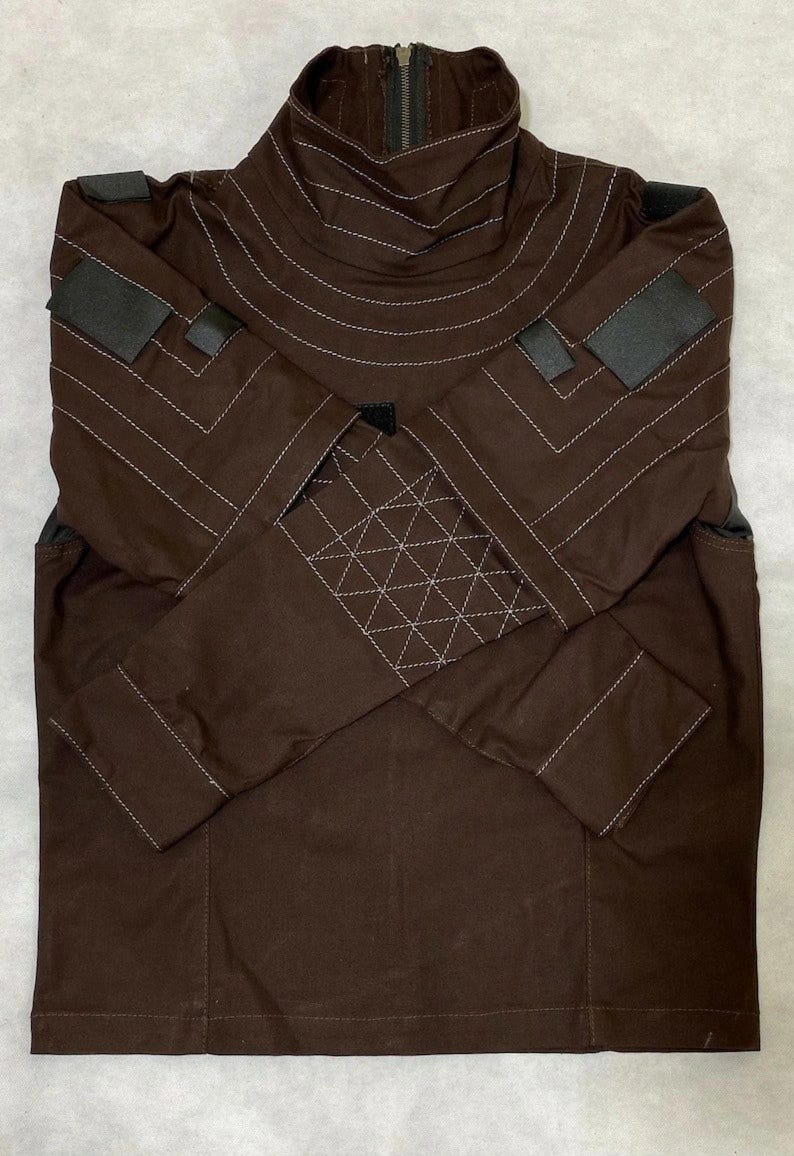 Puede incluir: Chaqueta marr&oacute;n con cuello alto y mangas largas. La chaqueta presenta detalles de costura blancos y parches negros en los hombros y las mangas. Una cremallera recorre la parte delantera del cuello.