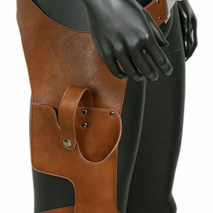 Op de afbeelding: Een bruine kunstlederen dijbeenholster met een brede riem met metalen gesp, een veiligheidsriem met drukknoopsluiting en een verstelbare beenband. De holster is afgewerkt met zichtbare stiksels en zilverkleurige metalen klinknagels.