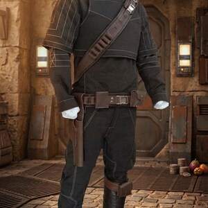 May include: A black costume with a high collar, short sleeves, and a brown leather strap across the chest. The costume includes matching trousers, a belt with a holster, and a brown leather accent on the leg. The outfit is displayed on a mannequin.