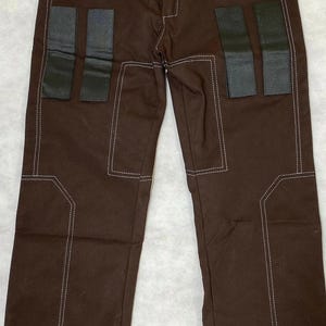 Puede incluir: Pantalones marrones con costuras blancas y parches rectangulares negros. Los pantalones tienen un dise&ntilde;o cl&aacute;sico con cierre de bot&oacute;n y cremallera. Los detalles de costura a&ntilde;aden un toque &uacute;nico.