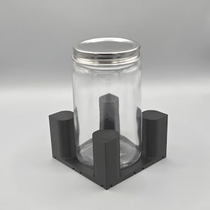 Könnte beinhalten: Ein klares Glas mit silbernem Deckel steht in einem dunkelgrauen, quadratischen Halter. Der Halter hat vier abgerundete Pfosten. Das Glas ist leer. Der Hintergrund ist einfarbig hellgrau.