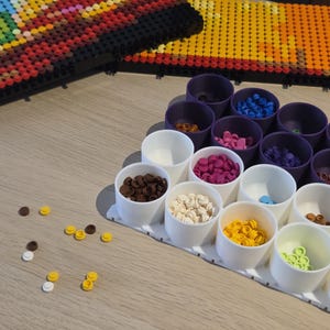 Könnte beinhalten: Eine Sammlung bunter Kunststoffperlen in verschiedenen Farben, darunter Braun, Weiß, Gelb und Rosa, ist in kleinen weißen und lila Behältern angeordnet. Die Perlen sind für ein Bastelprojekt bestimmt, mit größeren zusammengebauten Teilen im Hintergrund.