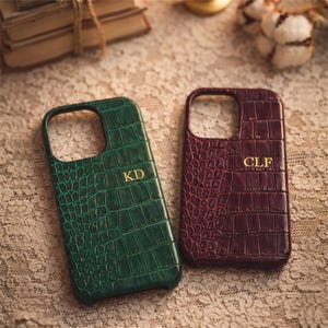 Puede incluir: Dos fundas para teléfono con textura de piel de cocodrilo. Una es verde oscuro con las iniciales "KD" en dorado. La otra es burdeos con las iniciales "CLF" en dorado. Ambas fundas tienen un recorte para la cámara y los botones.