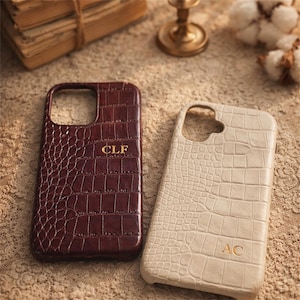 Könnte beinhalten: Zwei Handyhüllen mit Krokodilhautstruktur. Eine ist tief burgunderfarben mit den goldenen Buchstaben "CLF". Die andere ist cremefarben mit den goldenen Buchstaben "AC". Beide Hüllen haben eine rechteckige Form mit abgerundeten Ecken.