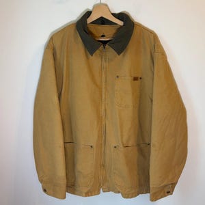 May include: A tan canvas work jacket featuring a dark olive green corduroy collar, a full-length front zipper, one chest pocket with a small rectangular label, and two large lower patch pockets with metal rivets at the corners.