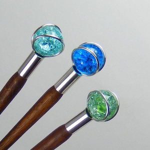 Könnte beinhalten: Drei dekorative Haarstäbe mit Holzgriff, silberfarbenem Metallakzent und einer Craquelé-Glaskugel in Blau- und Grüntönen. Die Kugeln sind in einem silberfarbenen Metallkäfig eingeschlossen.